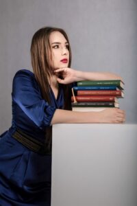 Anna Zborowska oparta o stos książek, granatowa sukienka, szare tło