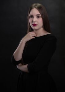 Anna Zborowska w czarnej sukience z dłonią przy szyi, ciemne tło