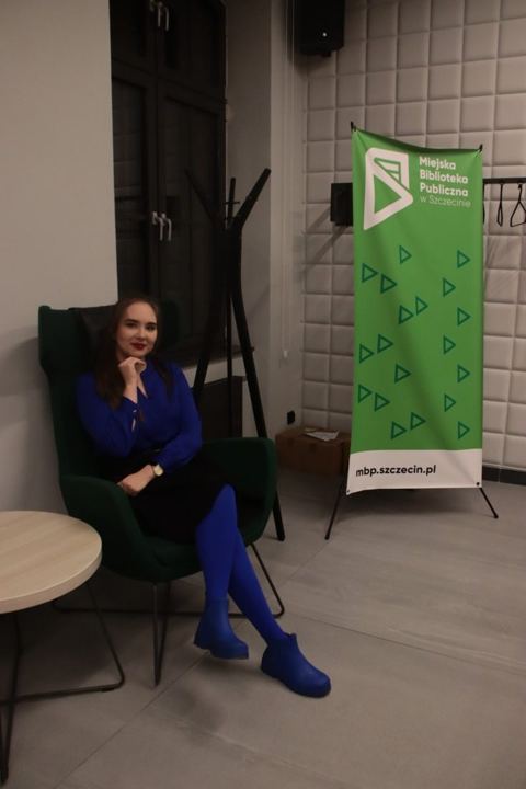 Anna Zborowska w fotelu obok banera biblioteki