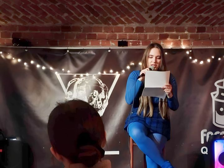 Anna Zborowska siedzi na stołku na scenie i czyta tekst z kartki, na pierwszym planie widz od tyłu